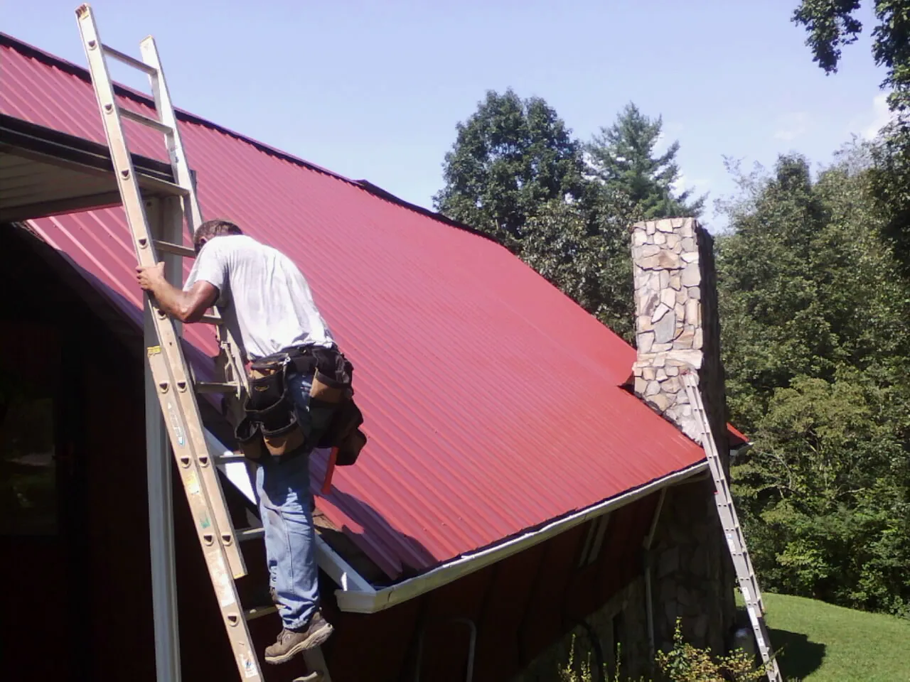 Roofer on a ladder beside a red metal roof and stone chimney — Soffit & Fascia Repair by Roofing Brothers in Tahlequah